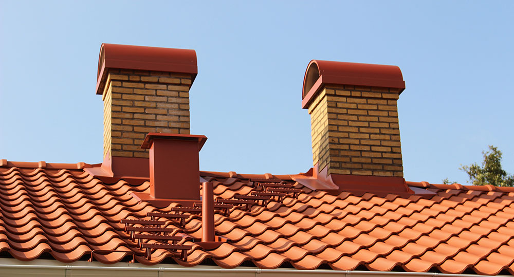 Två murade skorstenar och en ventilationshuv syns ovanför ett tak med röda takpannor mot en klar himmel.