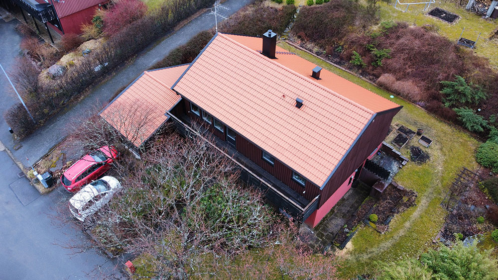 Flygfoto över ett mörkbrunt hus med rött tak, omgivet av träd och gräsmatta, med en uppfart där en röd bil är parkerad.