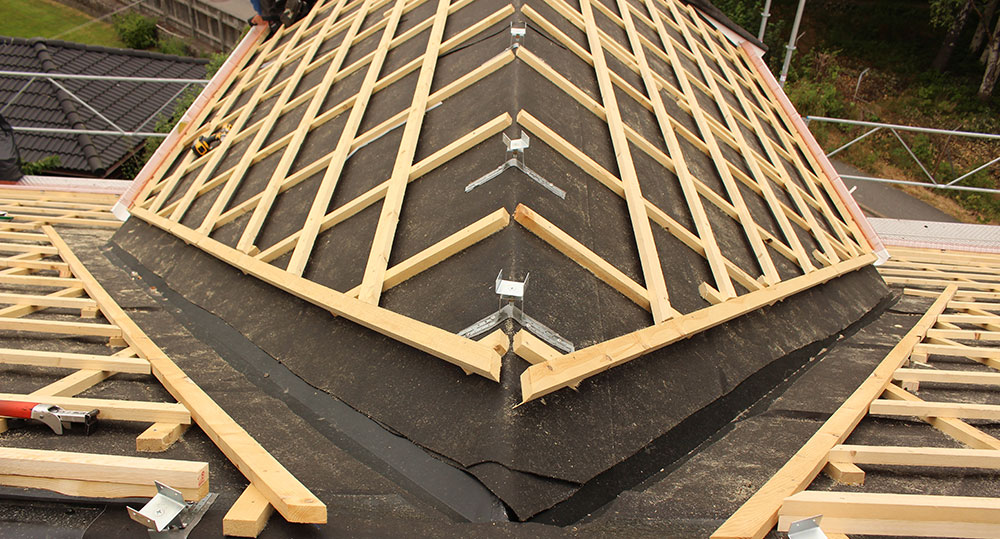 Närbild av ett tak under byggnation med synliga träreglar, takpapp och beslag vid nocken.